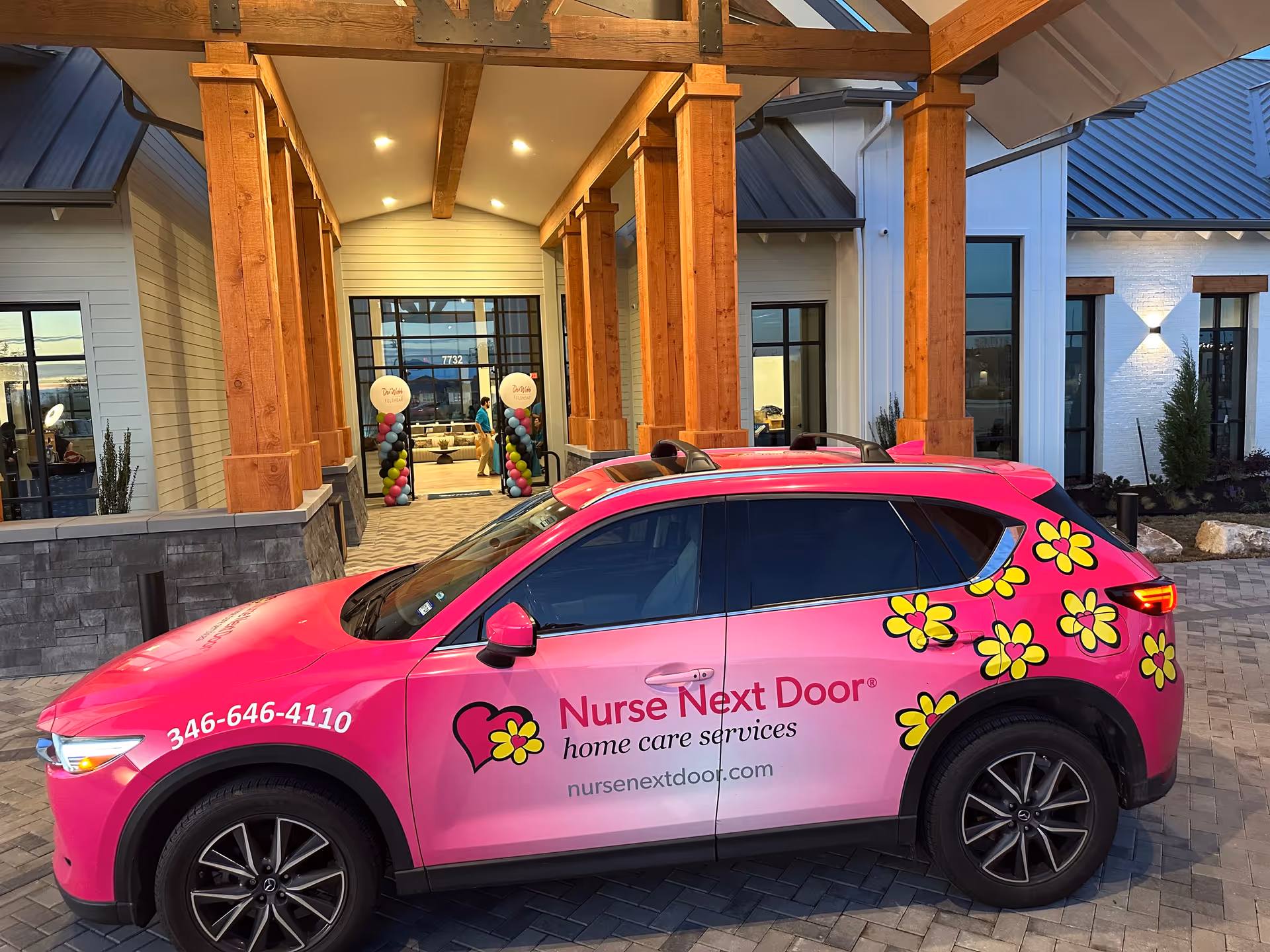 Pink Nurse Next Door vehicle parked outside a modern facility entrance.