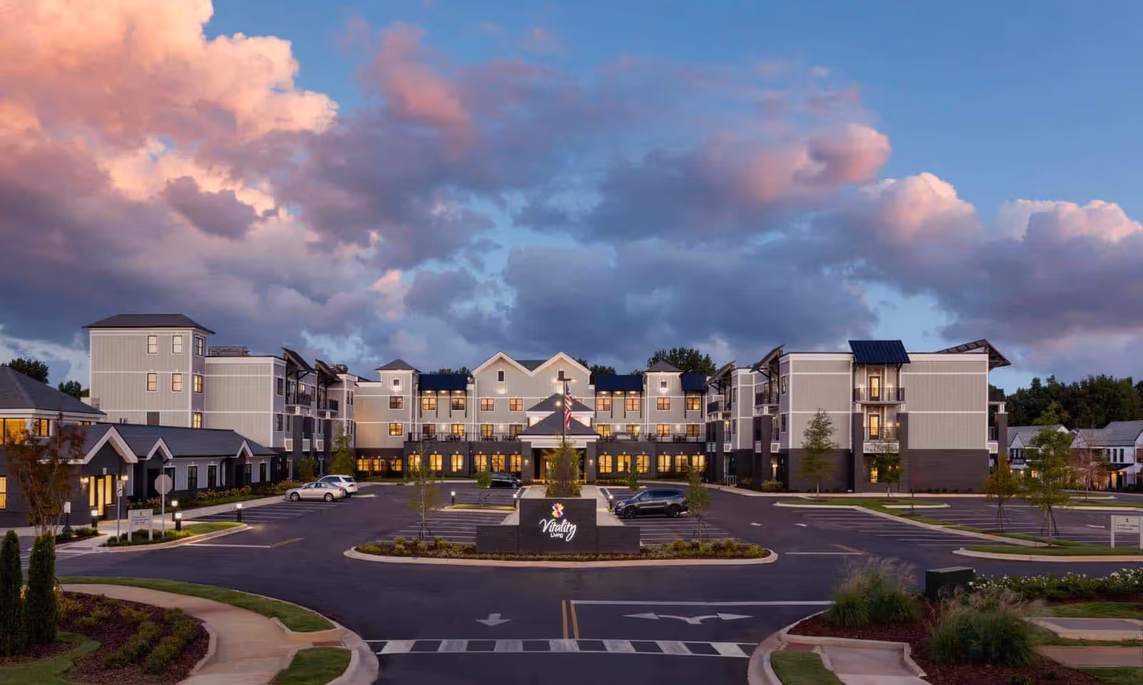 Exterior front view of a multi-story senior living facility at dusk with a circular driveway, parked cars, and illuminated windows under dramatic clouds.