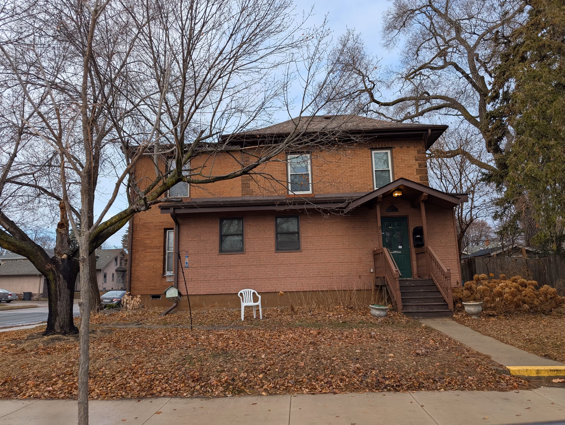 Two-story brick house with a covered front porch and green door, leafless trees and a white plastic chair on a leaf-covered lawn.