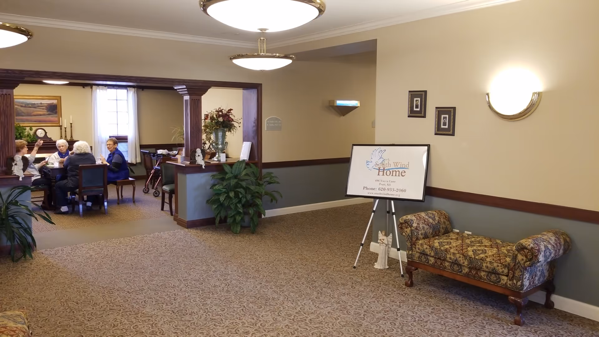 Indoor lobby with a seating area, signboard, plants, and a group of elders at a table.