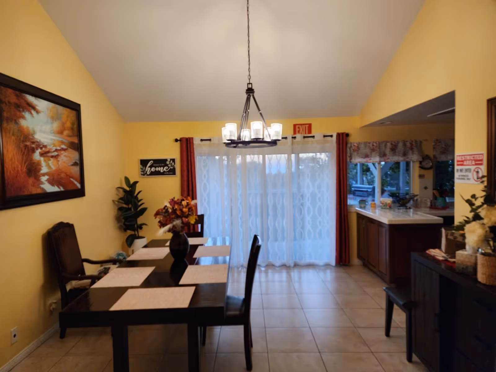 A dining room with a dark wooden table set with six beige placemats and a vase of flowers in the center. There are six chairs around the table. The walls are painted yellow, and a large painting of a nature scene hangs on the left wall. A chandelier with multiple lights hangs above the table. At the back, sheer white curtains cover a sliding glass door with red drapes on either side. To the right, there is a kitchen counter with various items on it and a window with floral valances. The floor is tiled, and there are decorative plants and signs on the walls.