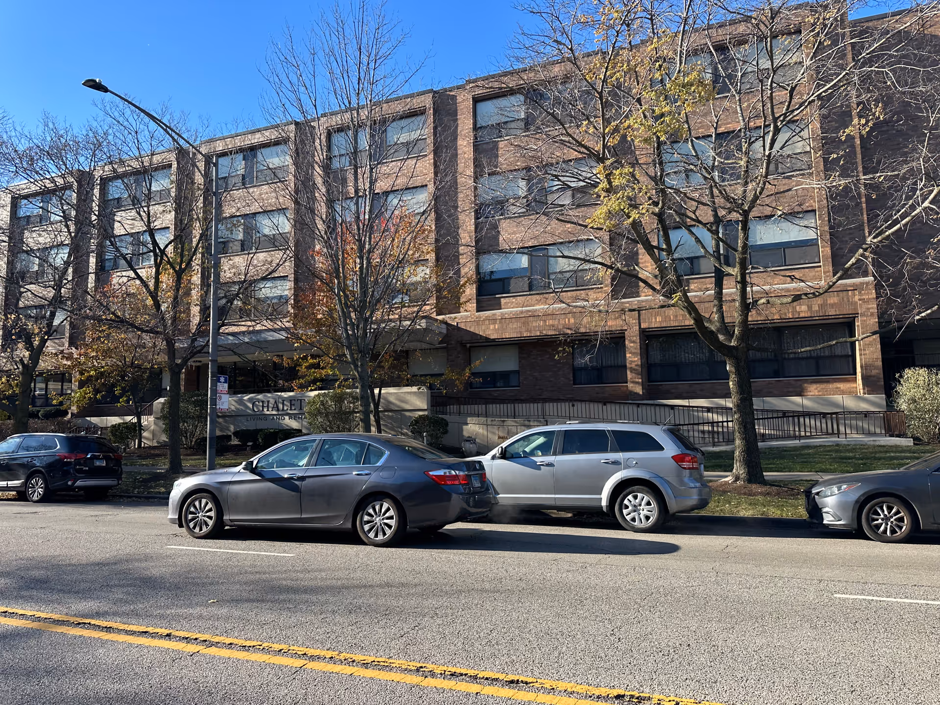 Exterior view of a multi-story brick building with several windows, identified as Chalet Living And Rehab Center. There are three cars parked on the street in front of the building, and leafless trees line the sidewalk. The sky is clear and blue.