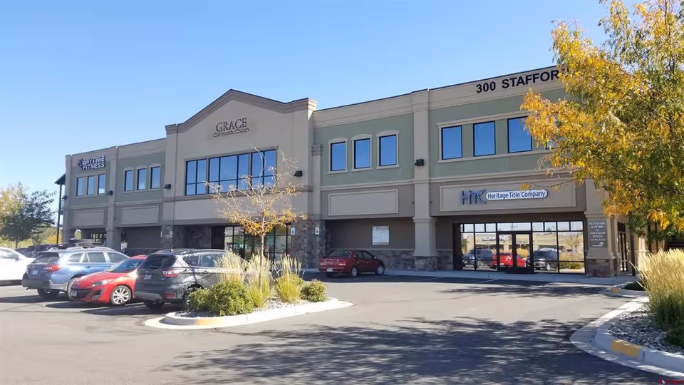 Two-story commercial building with multiple storefronts, a parking lot, trees, and blue sky.