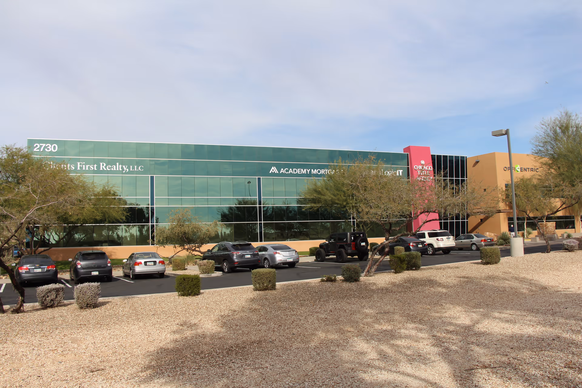 Exterior view of a modern glass-front building with a parking lot and desert landscaping.
