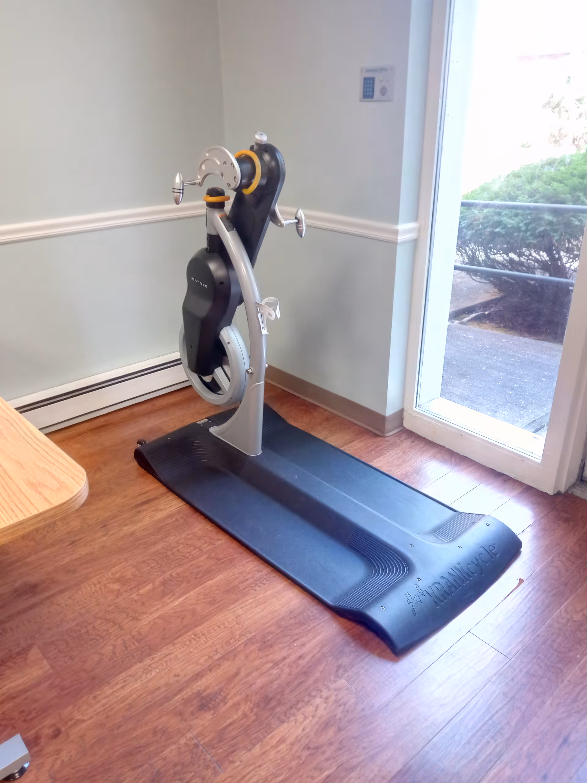 Recumbent exercise machine on a wood floor beside a glass door in a senior living facility.