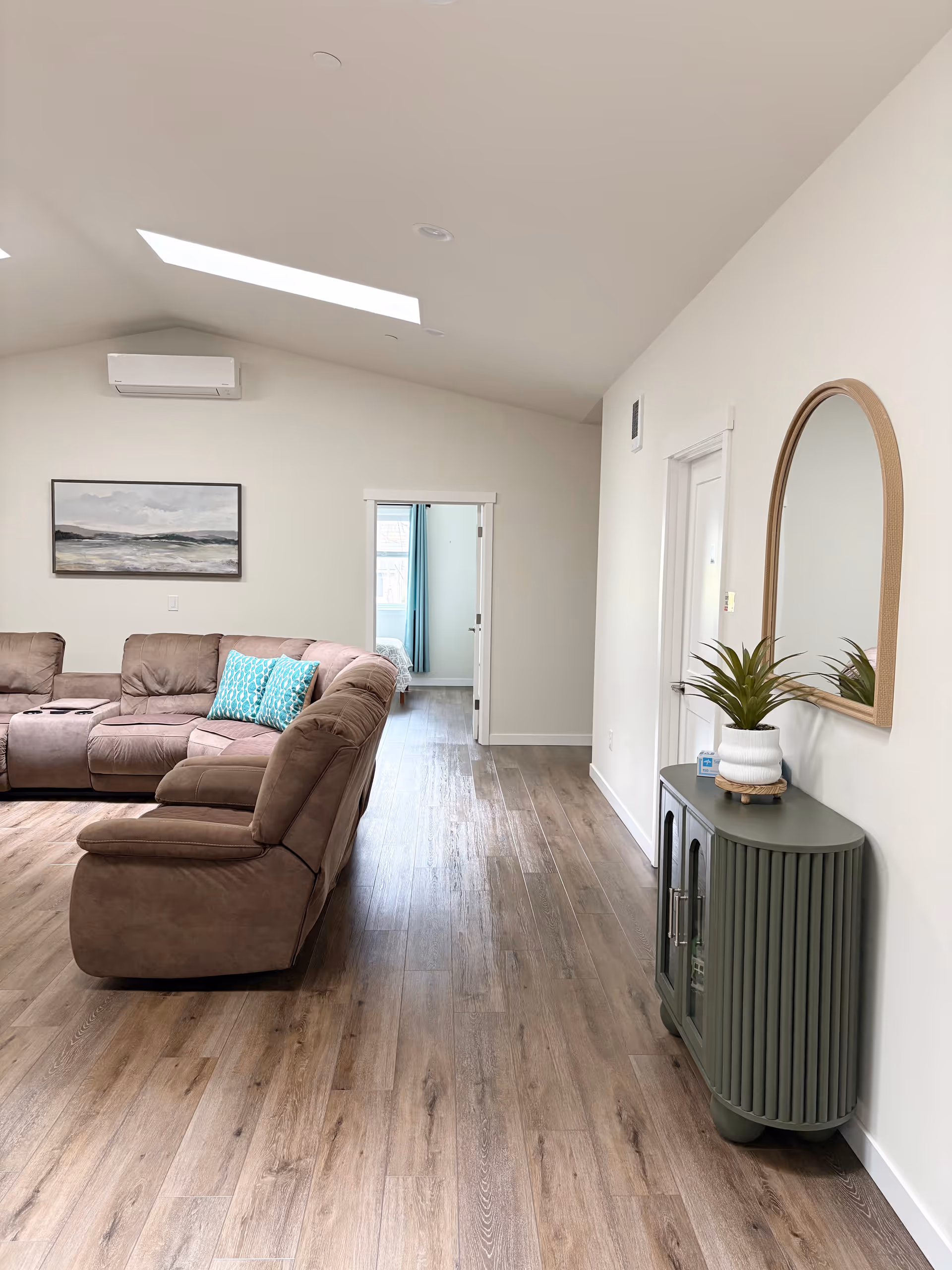 Bright living room with brown sectional, green console, mirror, and doorway to a bedroom.
