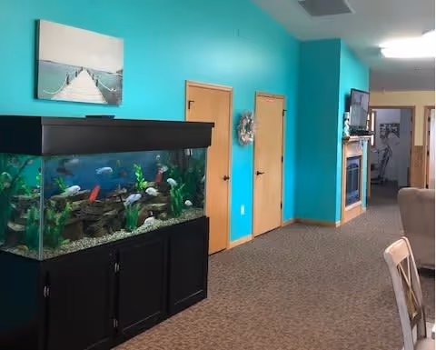 Interior view of a senior living facility hallway with turquoise walls, a large fish tank on a black stand, two wooden doors, a wall-mounted TV above a fireplace, and part of a dining table and chair visible on the right.