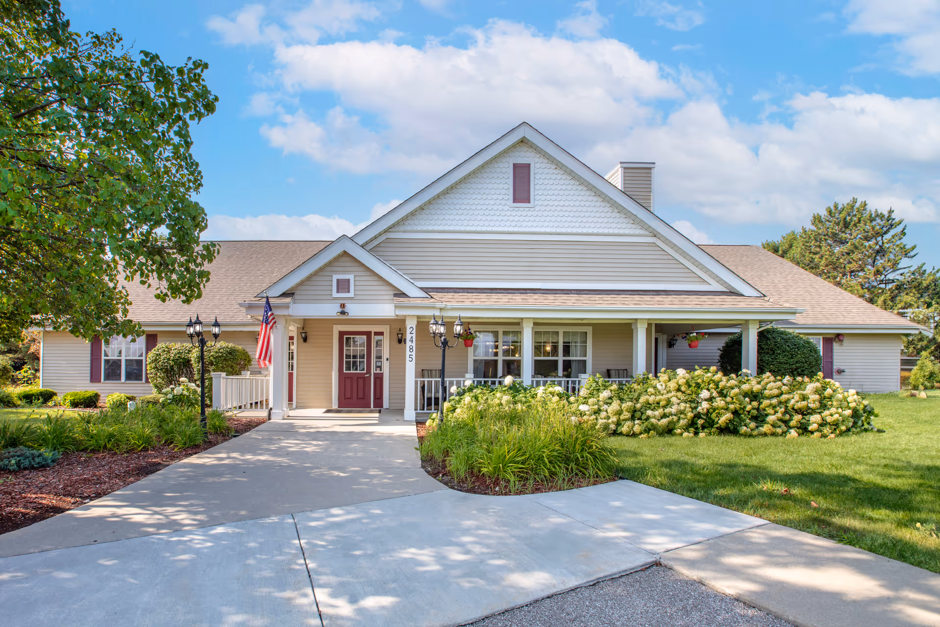 Front exterior view of a single-story building with beige siding and a gabled roof. The entrance features a covered porch with white columns, a red door, and an American flag. There are well-maintained shrubs, flowering plants, and a tree on the left side. The sky is partly cloudy with blue patches.