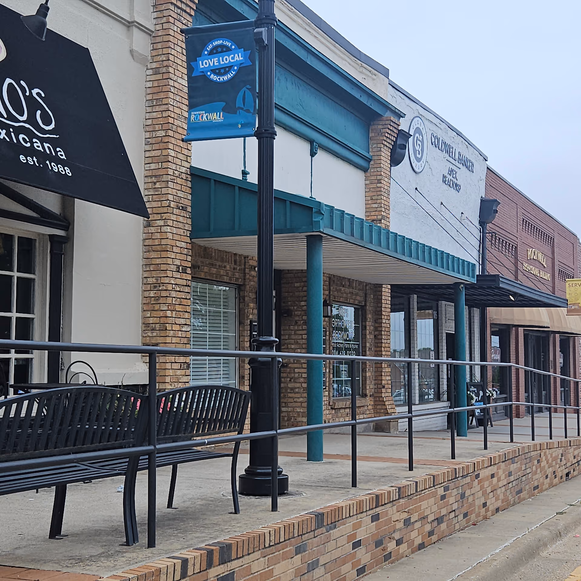 Row of storefronts with brick facades, awnings, benches, and a lamppost along the sidewalk.
