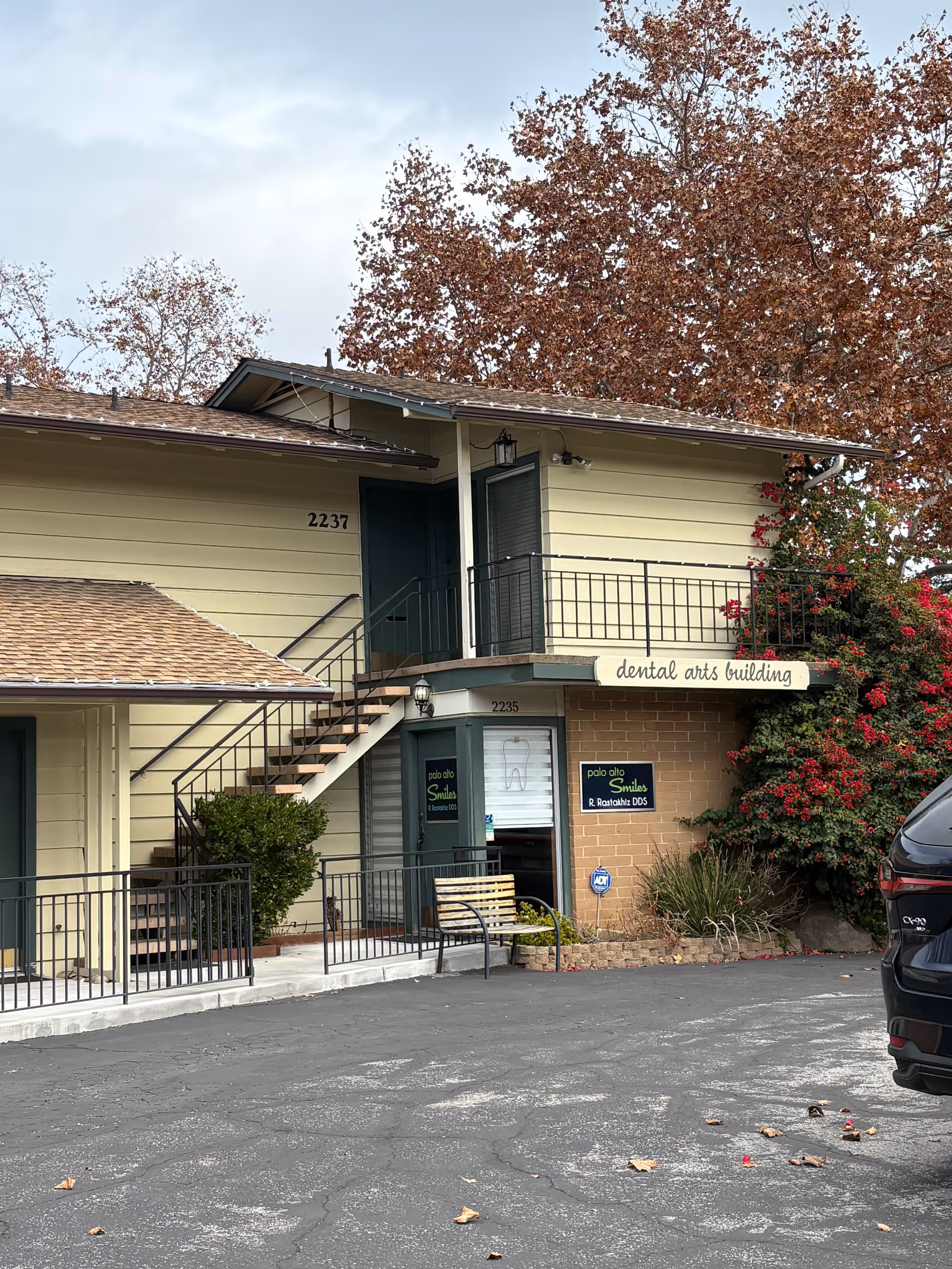 Two-story beige building with exterior stairs, signs, potted plants, and a parked car outside.