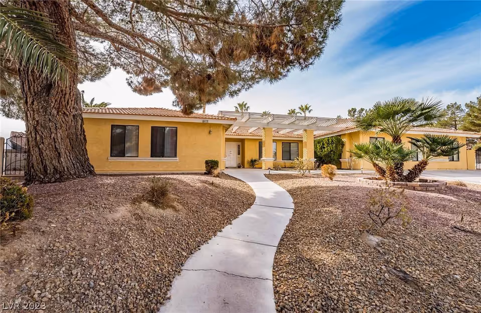 Single-story yellow stucco building with a curved walkway, desert landscaping, and a pergola-covered entrance.