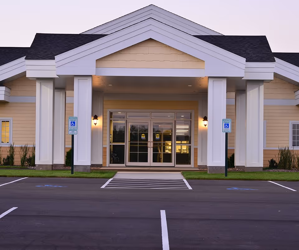 Covered front entrance of a senior living facility with white columns, glass double doors, and marked accessible parking spaces.