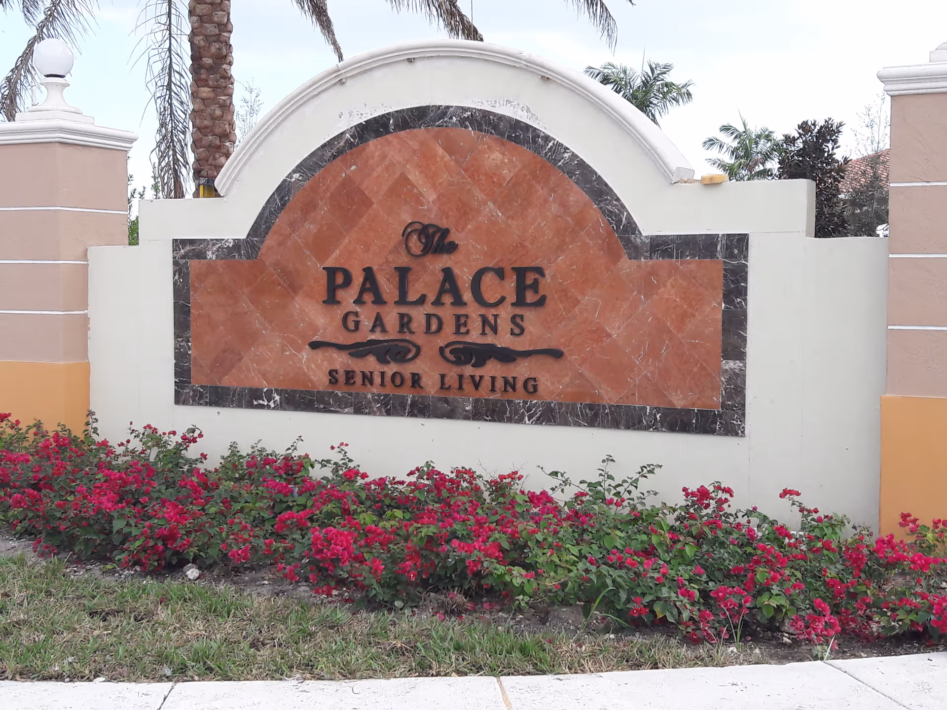 Front entrance sign reads The Palace Gardens Senior Living, surrounded by pink flowers.