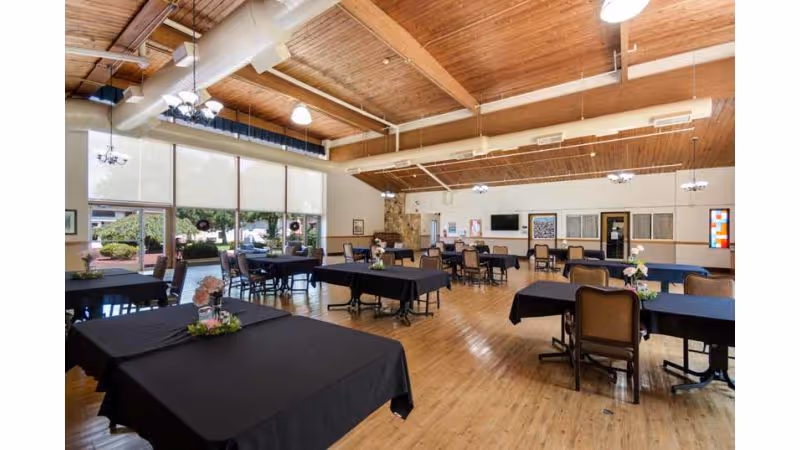 Spacious dining room with wooden floors and ceiling beams, several tables covered with black tablecloths, each decorated with small floral centerpieces. Large windows with roller shades let in natural light, and there are multiple chairs around the tables. The room has a cozy and inviting atmosphere with a stone fireplace and wall-mounted TV in the background.