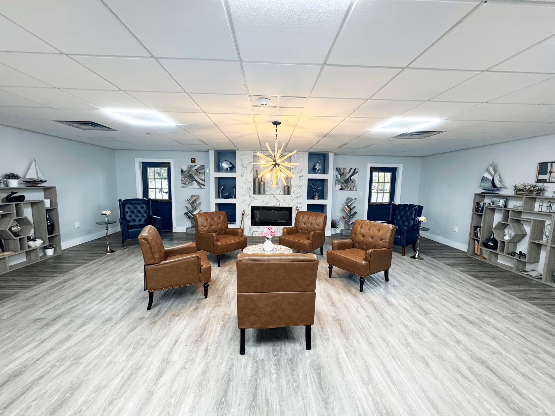 A spacious and modern living room with light wood flooring and a white ceiling with recessed lighting. The room features a central seating area with five brown leather armchairs arranged around a small round table with a vase of pink flowers. On either side of the room, there are blue armchairs near dark blue doors with windows. The back wall has a decorative fireplace surrounded by white and blue shelving units displaying various decorative items and artwork. Two wooden shelving units with nautical-themed decor are positioned against the side walls.