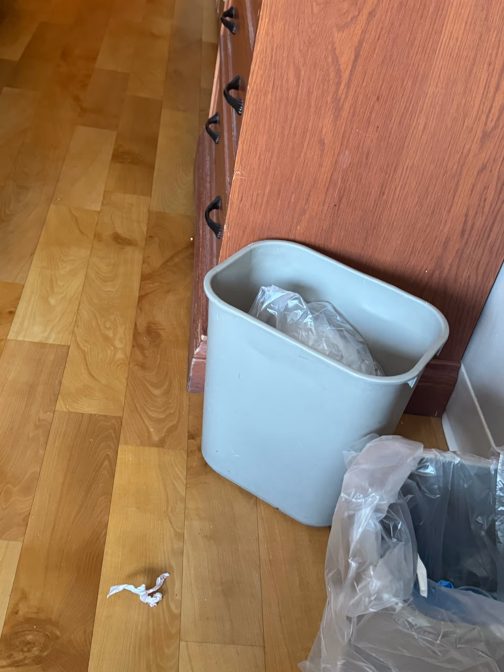 Blue trash can with a plastic bag beside wooden cabinets on a wooden floor.