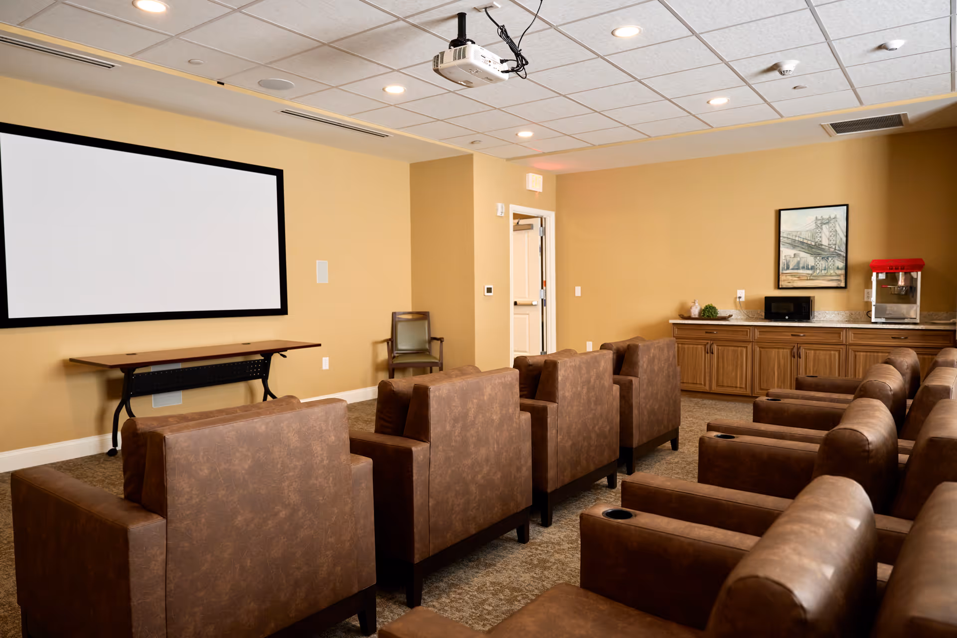 Senior living lounge with brown theater-style chairs facing a large projector screen.