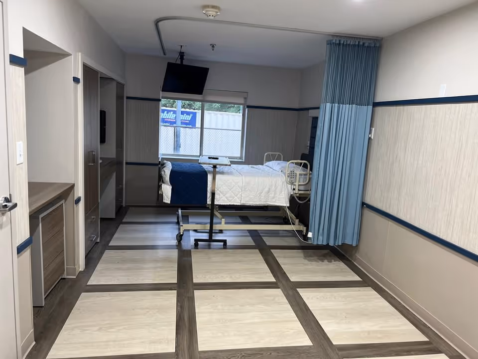 A clean and modern patient room in a care facility featuring a hospital bed with white and blue bedding, a mounted flat-screen TV, a privacy curtain, a window with a view outside, and built-in cabinetry along the left wall.