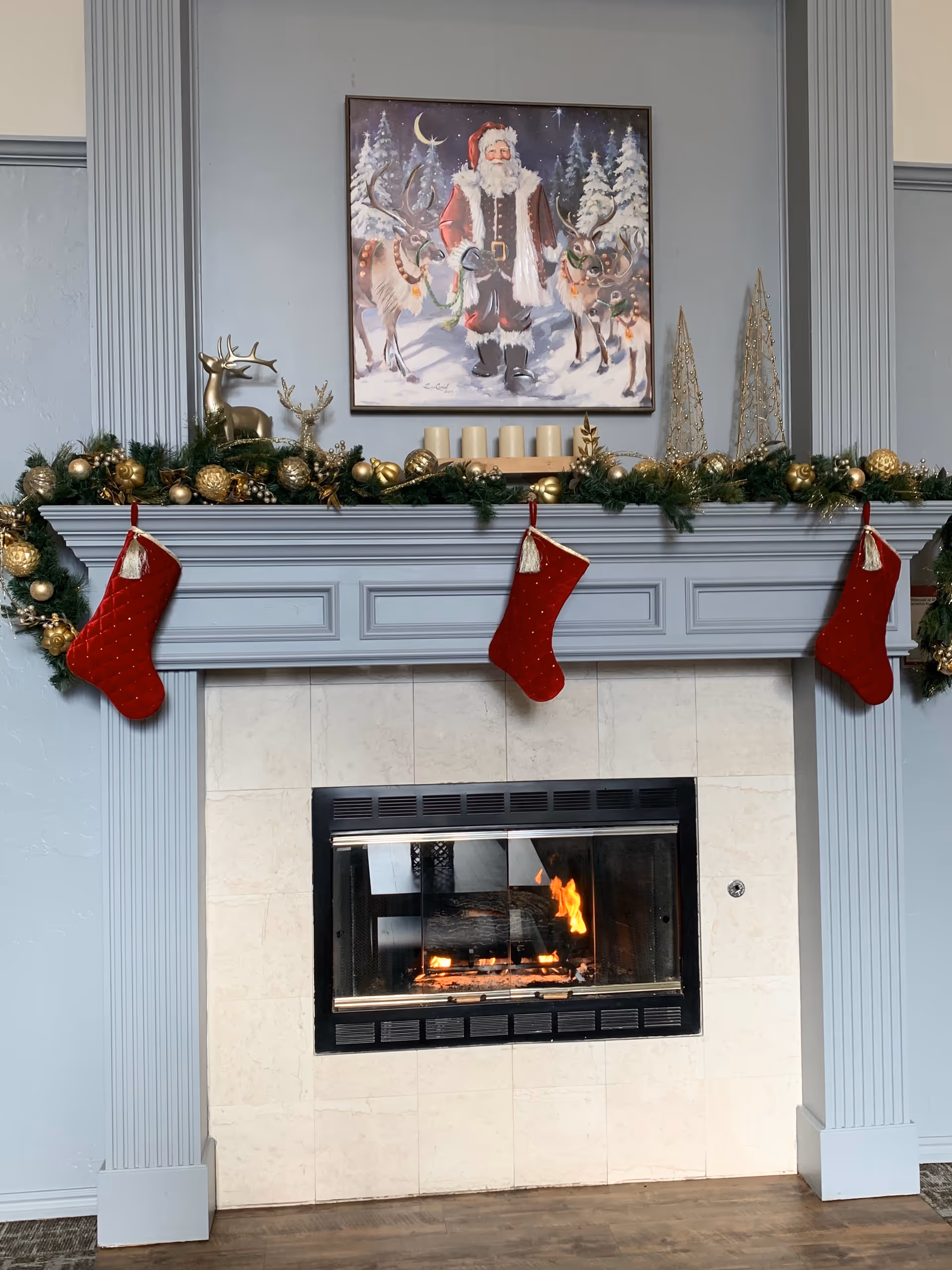 A decorated fireplace mantel with garland, stockings, candles, and Santa painting in a cozy interior.