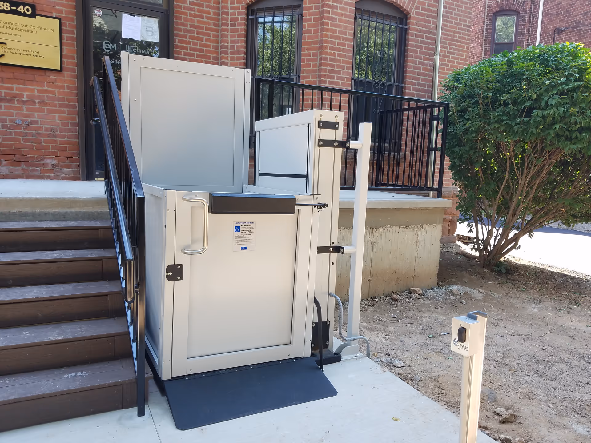 An outdoor wheelchair lift beside a brick building entrance with stairs and surrounding greenery.