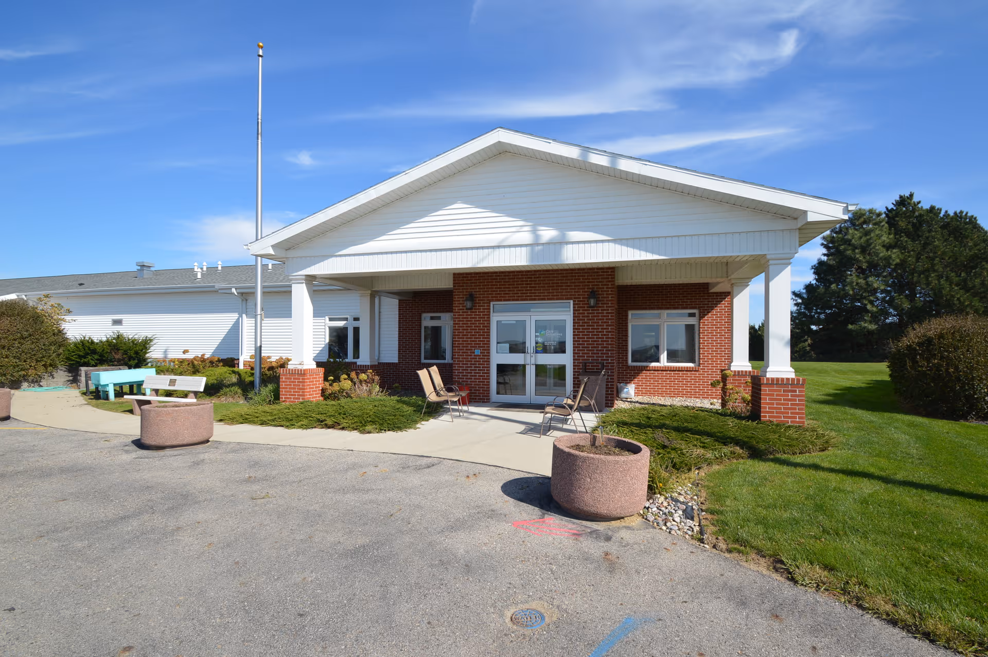 Front exterior view of Dunlap Independent Living facility with a covered entrance, brick and white siding walls, two chairs near the entrance, a flagpole, benches, and landscaped greenery under a clear blue sky.