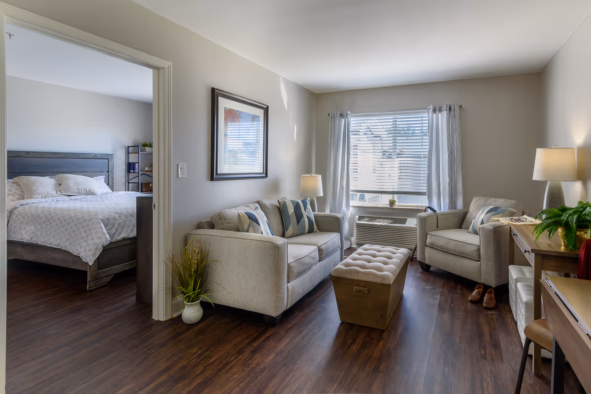 Bright living room with two beige sofas, ottoman, window, desk, and doorway to a bedroom.