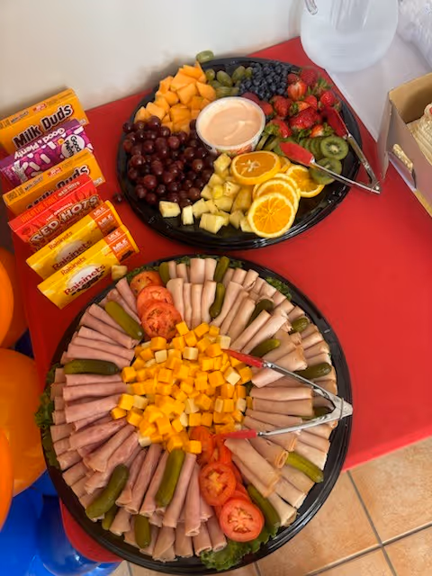 Two platters of deli meats, cheese, fruit, and vegetables on a red table.