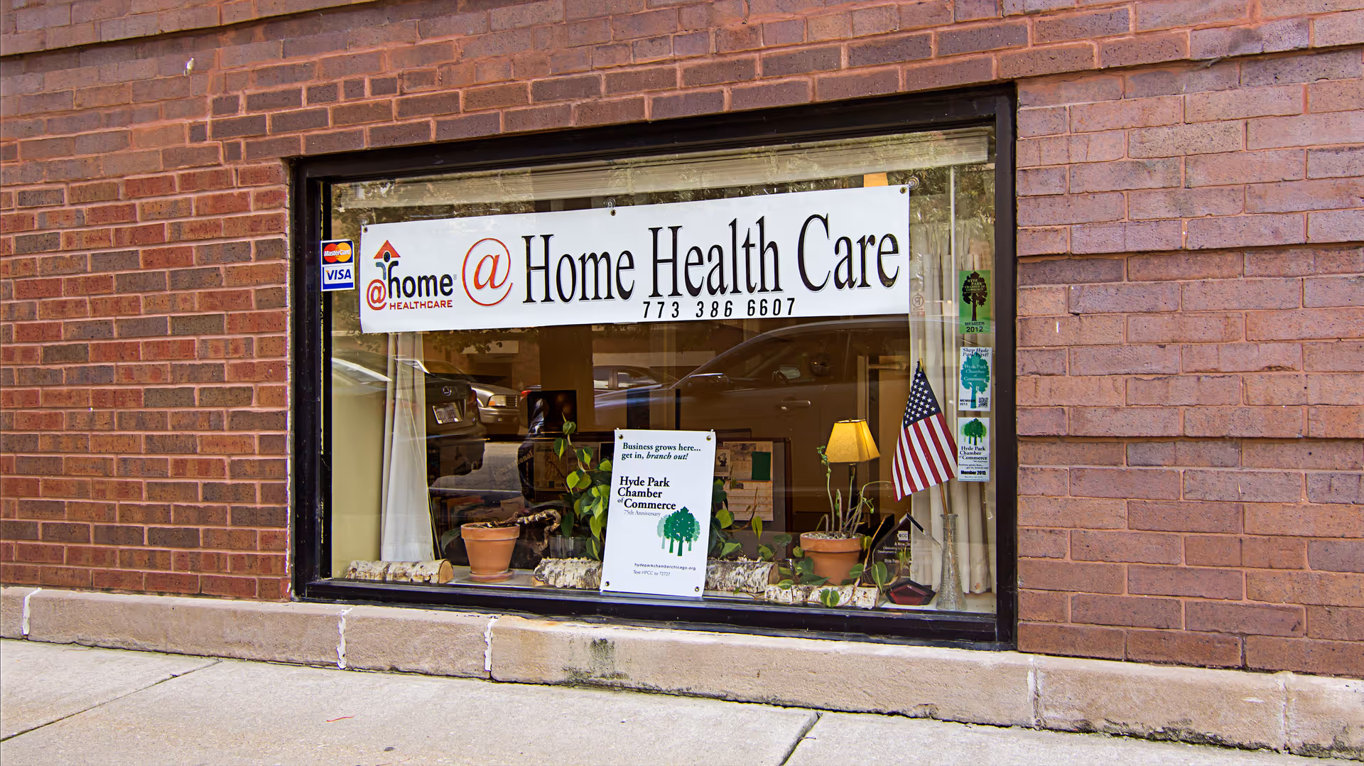 Brick storefront window displays @ Home Health Care sign with plants and flag.