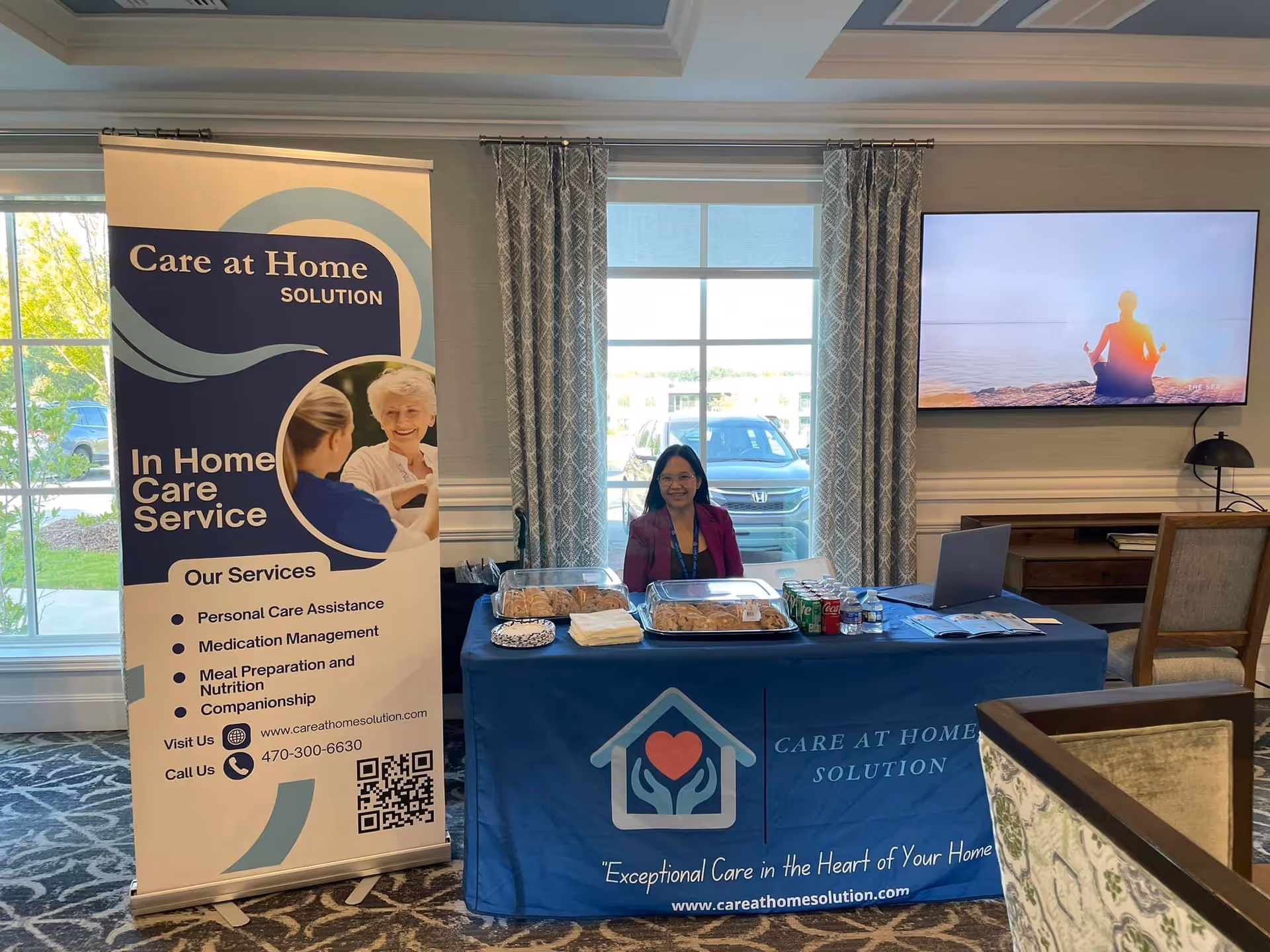 Indoor booth setup for Care at Home Solution with banner, table, staff, and TV.