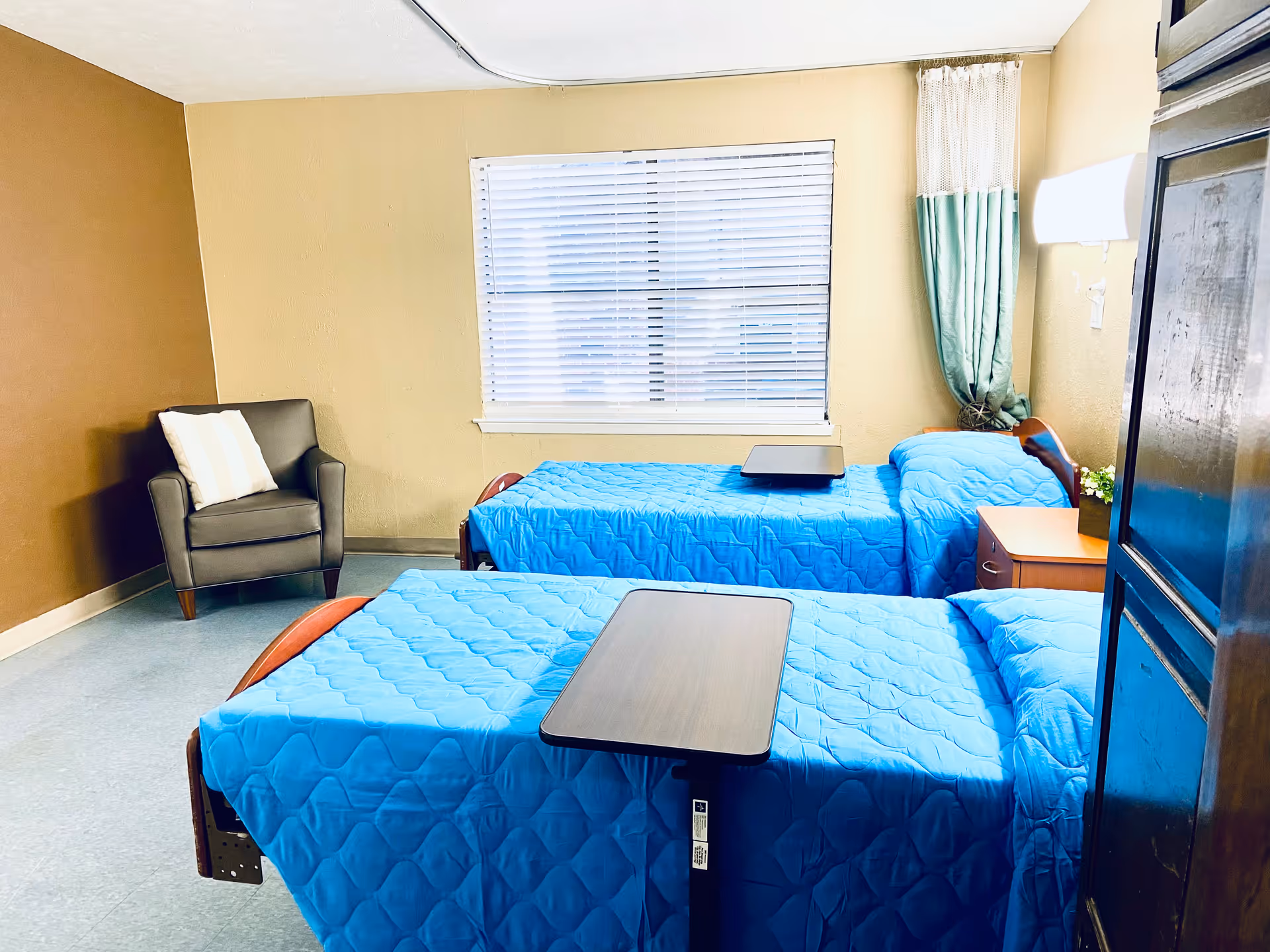 A bedroom with two single beds covered in bright blue quilts, each with an overbed table. There is a window with white blinds and a curtain on the right side. A gray armchair with a white pillow is placed in the corner against a brown wall. A small wooden nightstand with a plant is next to one of the beds.