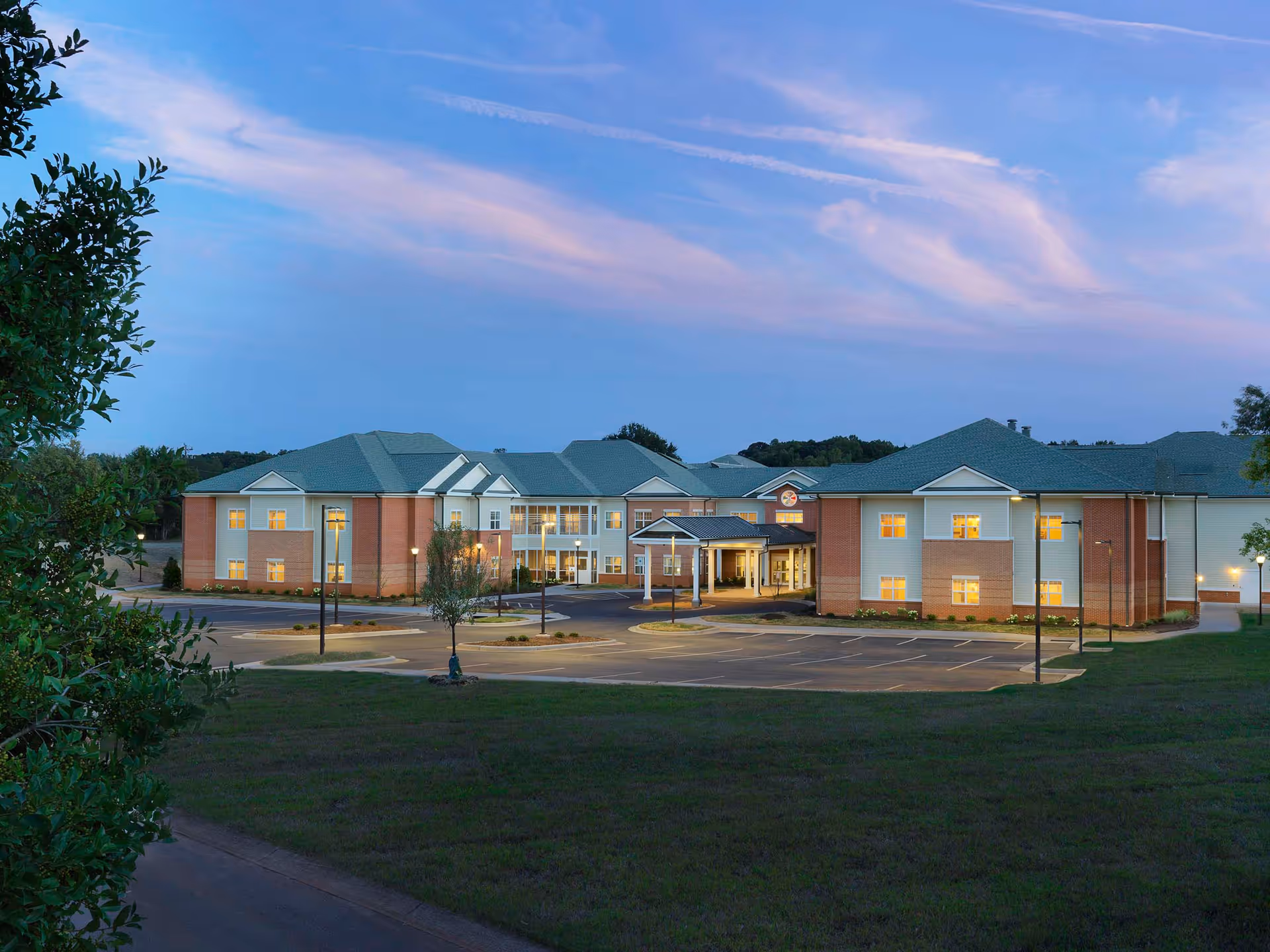 Exterior front view of a two-story senior living facility with lit windows, a covered entrance and adjacent parking lot at dusk.