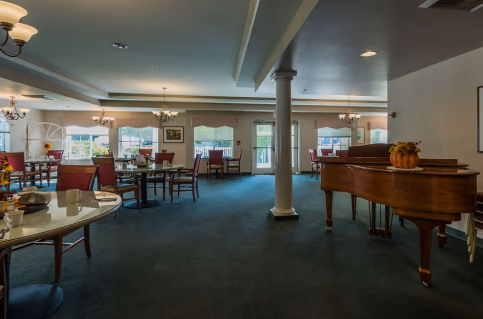 Spacious dining room with round tables and red chairs, a grand piano, and large windows letting in natural light.