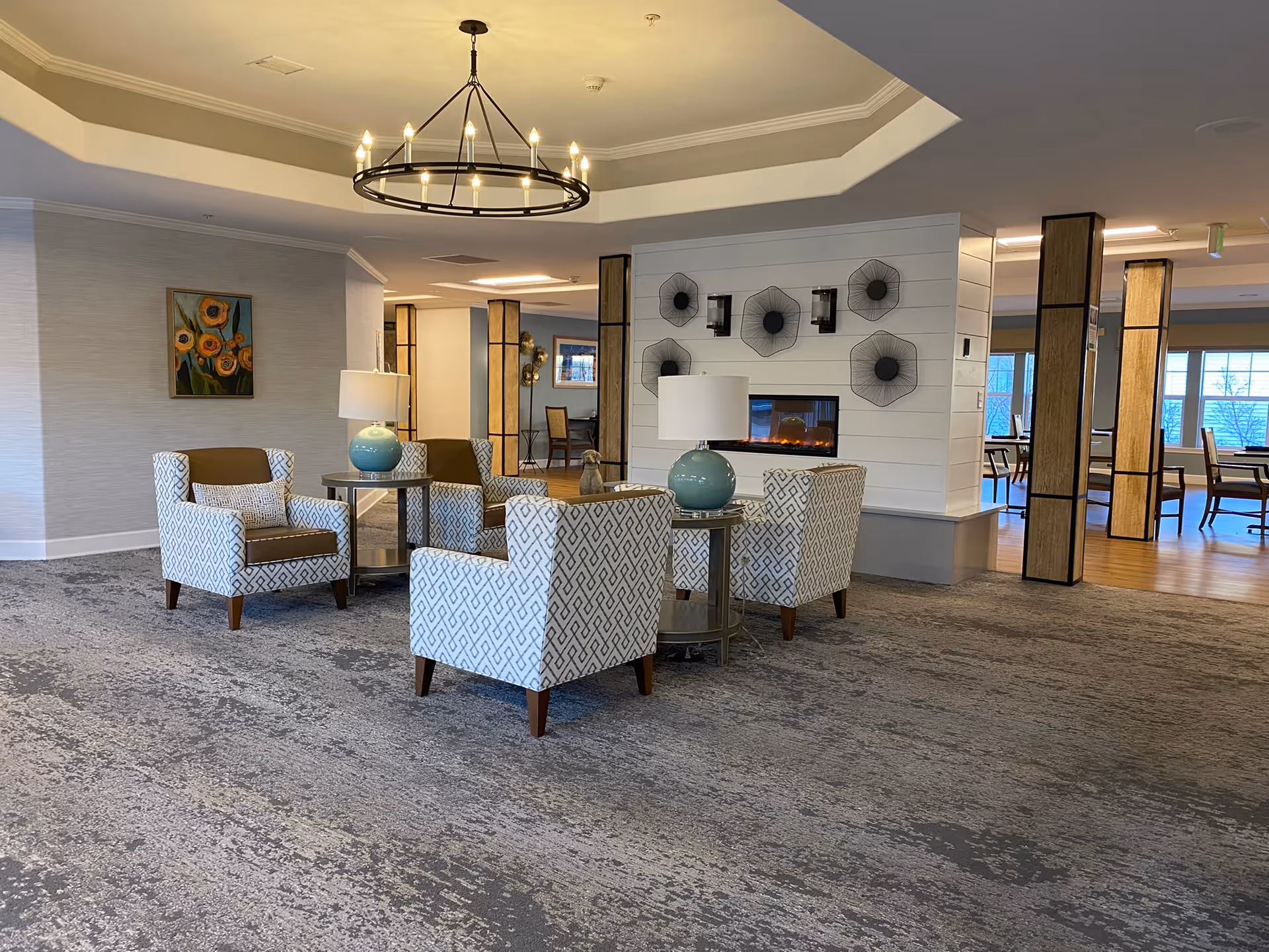 Spacious senior living lounge with patterned armchairs arranged around side tables, decorative lamps, a modern chandelier, and a fireplace wall.
