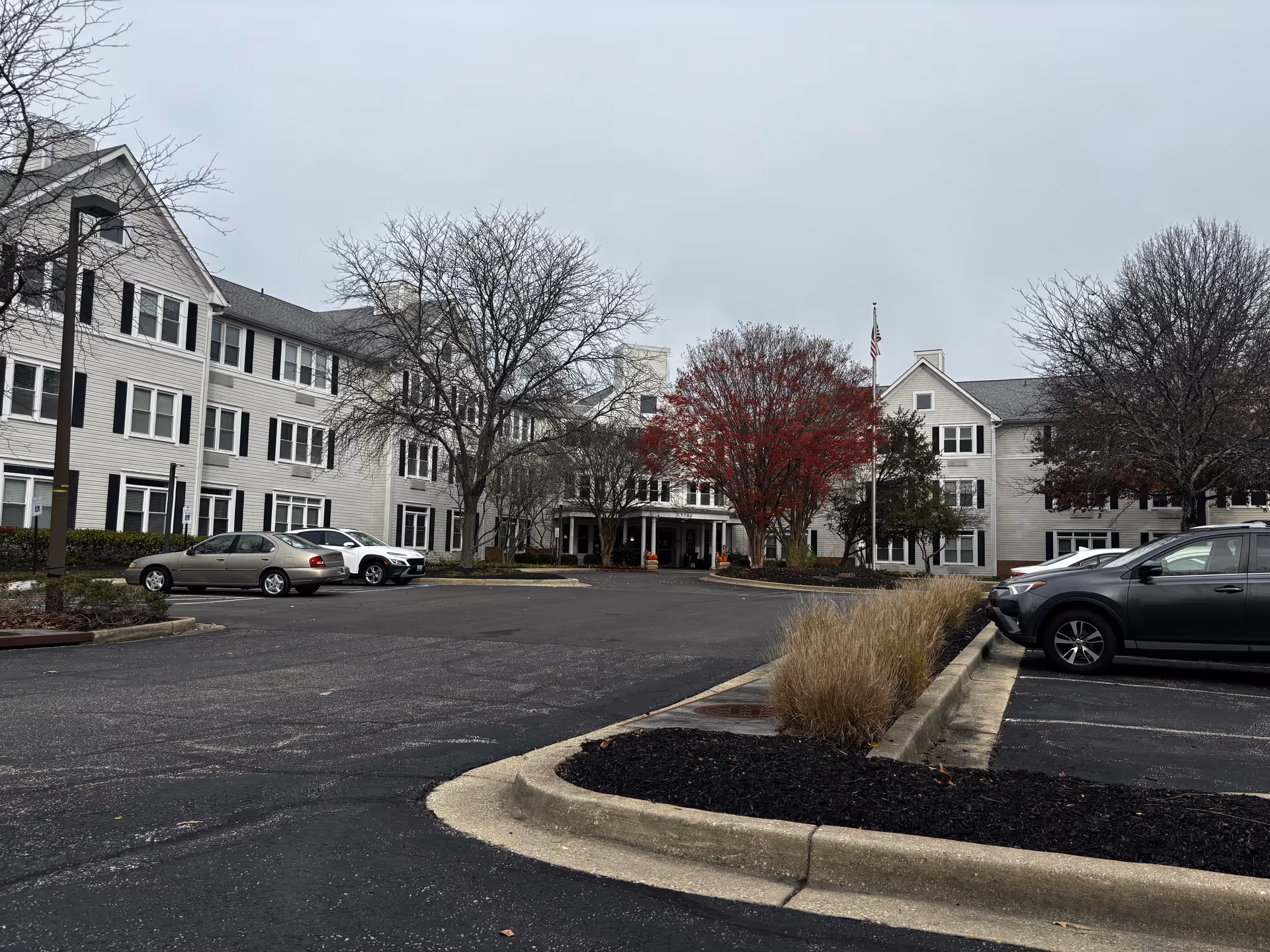 Exterior view of Morningstar Assisted Living facility showing a large three-story building with white siding and black shutters. There are several leafless trees and one tree with red leaves in front of the building. A parking lot with several parked cars is visible in the foreground under an overcast sky.