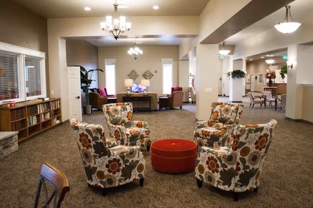 Cozy senior living lounge with floral chairs around a red ottoman, lamps, and bookshelves.