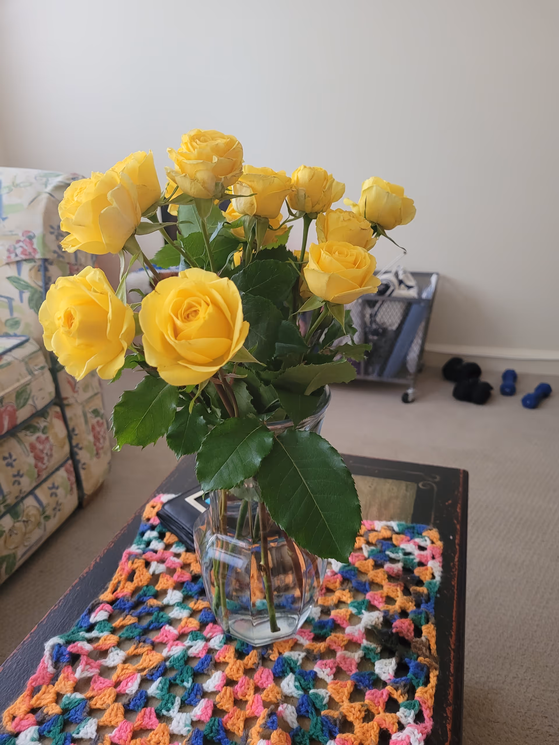 A vase of yellow roses on a colorful crocheted mat atop a coffee table.