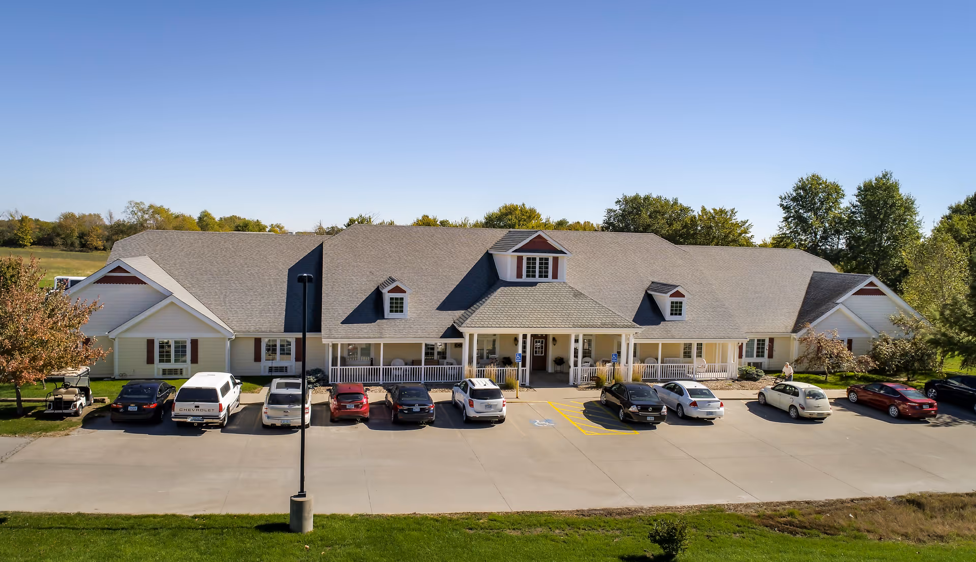 Front exterior view of Homestead Assisted Living of Chariton, a single-story building with a large porch, multiple dormer windows, and a parking lot with several cars parked in front. The building is surrounded by green grass and trees under a clear blue sky.