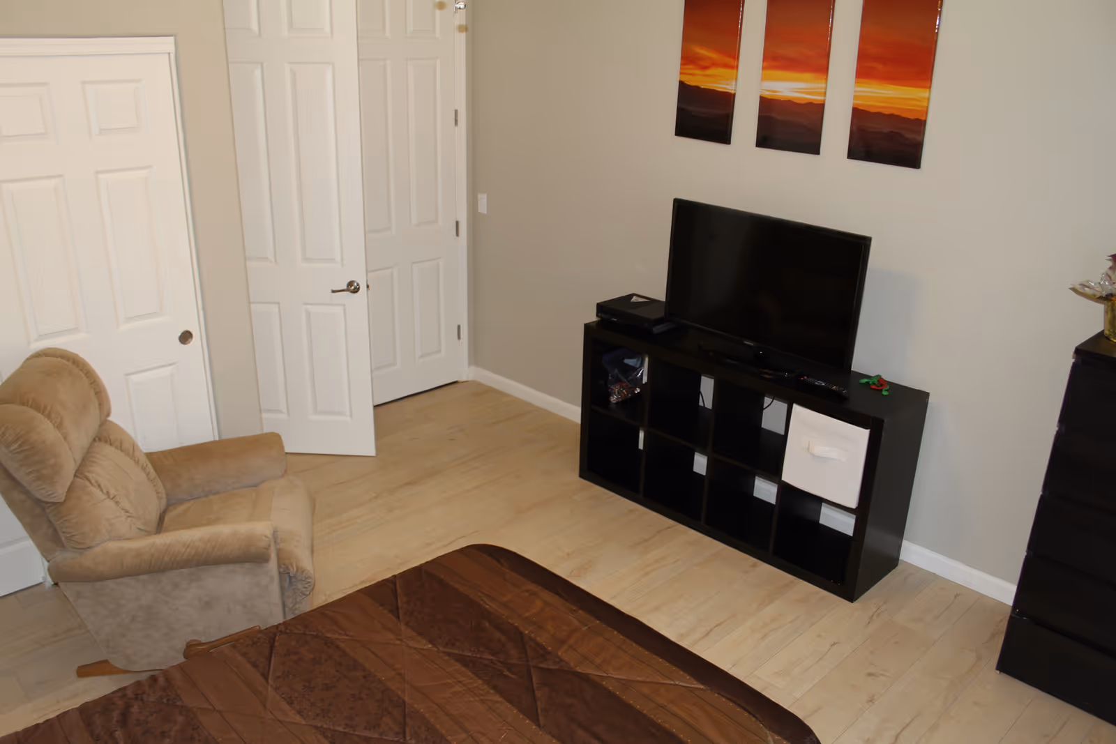 A bedroom with light wood flooring, a brown recliner chair, a bed with a brown quilt, a black TV stand with cubby holes holding a flat-screen TV and some items, a black dresser, and a three-panel sunset landscape artwork on the wall.