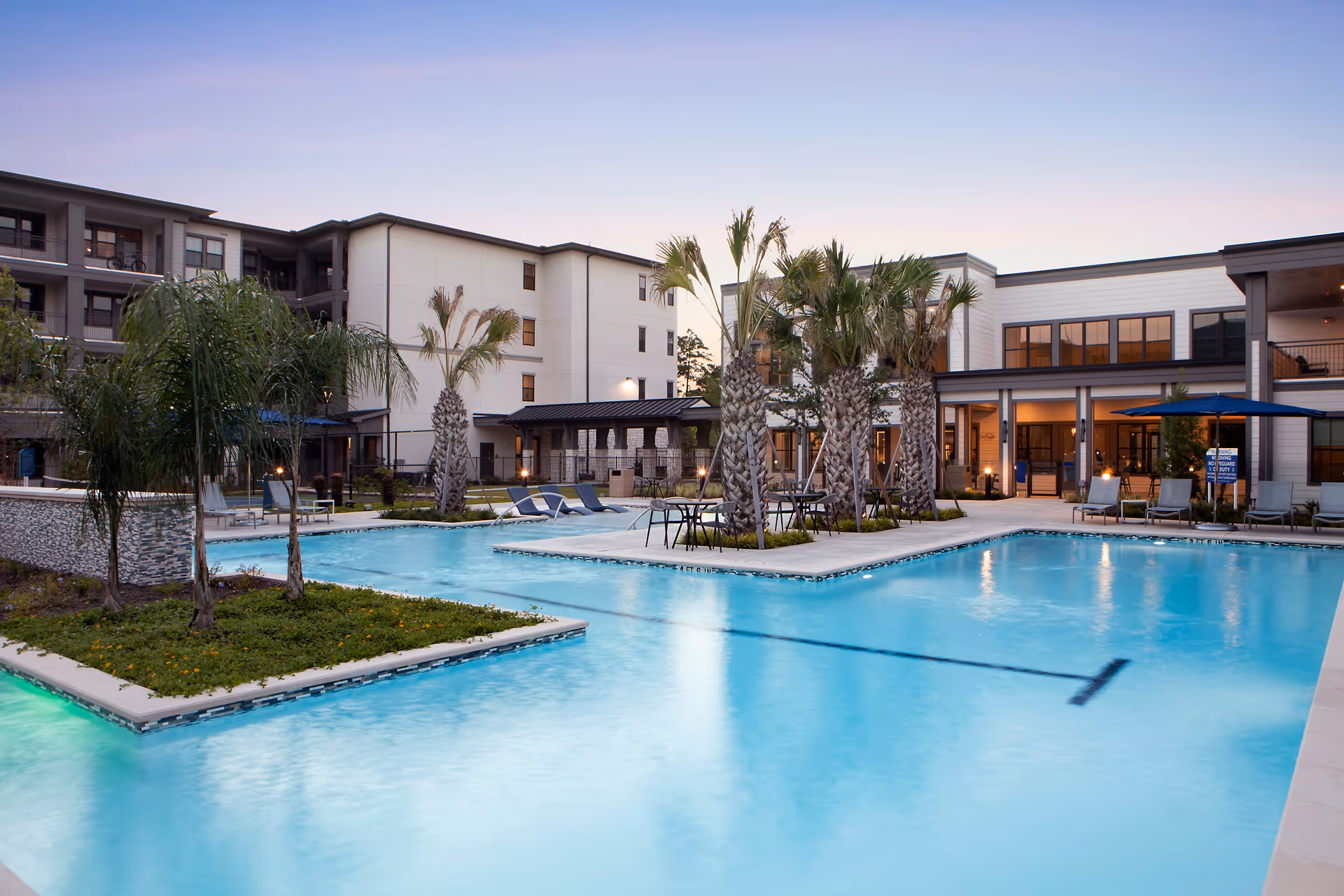 Outdoor swimming pool area at Mera Vintage Park with palm trees, lounge chairs, tables with umbrellas, and a multi-story building in the background during dusk.