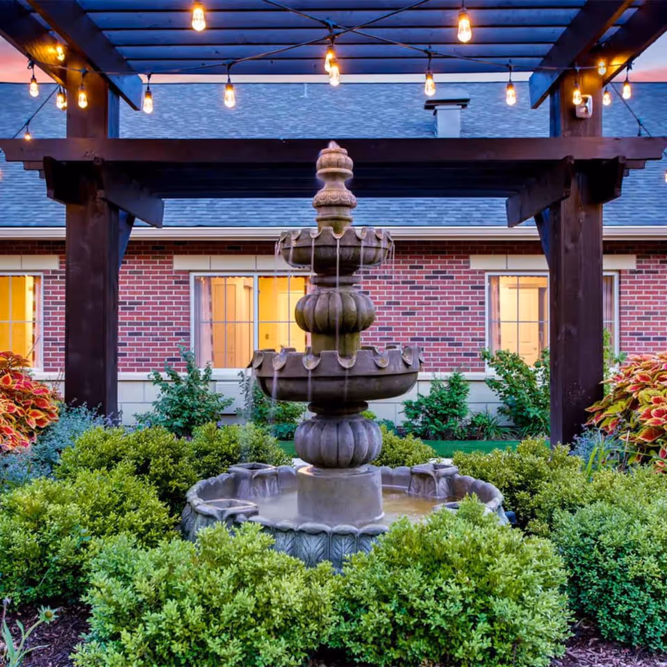 A tiered stone water fountain surrounded by green bushes and colorful plants under a wooden pergola with hanging string lights. In the background, there is a brick building with windows illuminated from inside.