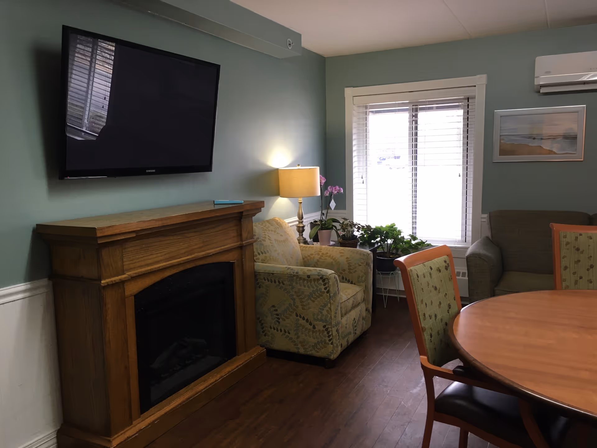 Living room in a senior facility with fireplace, wall-mounted TV, chairs, plants, and a round table.