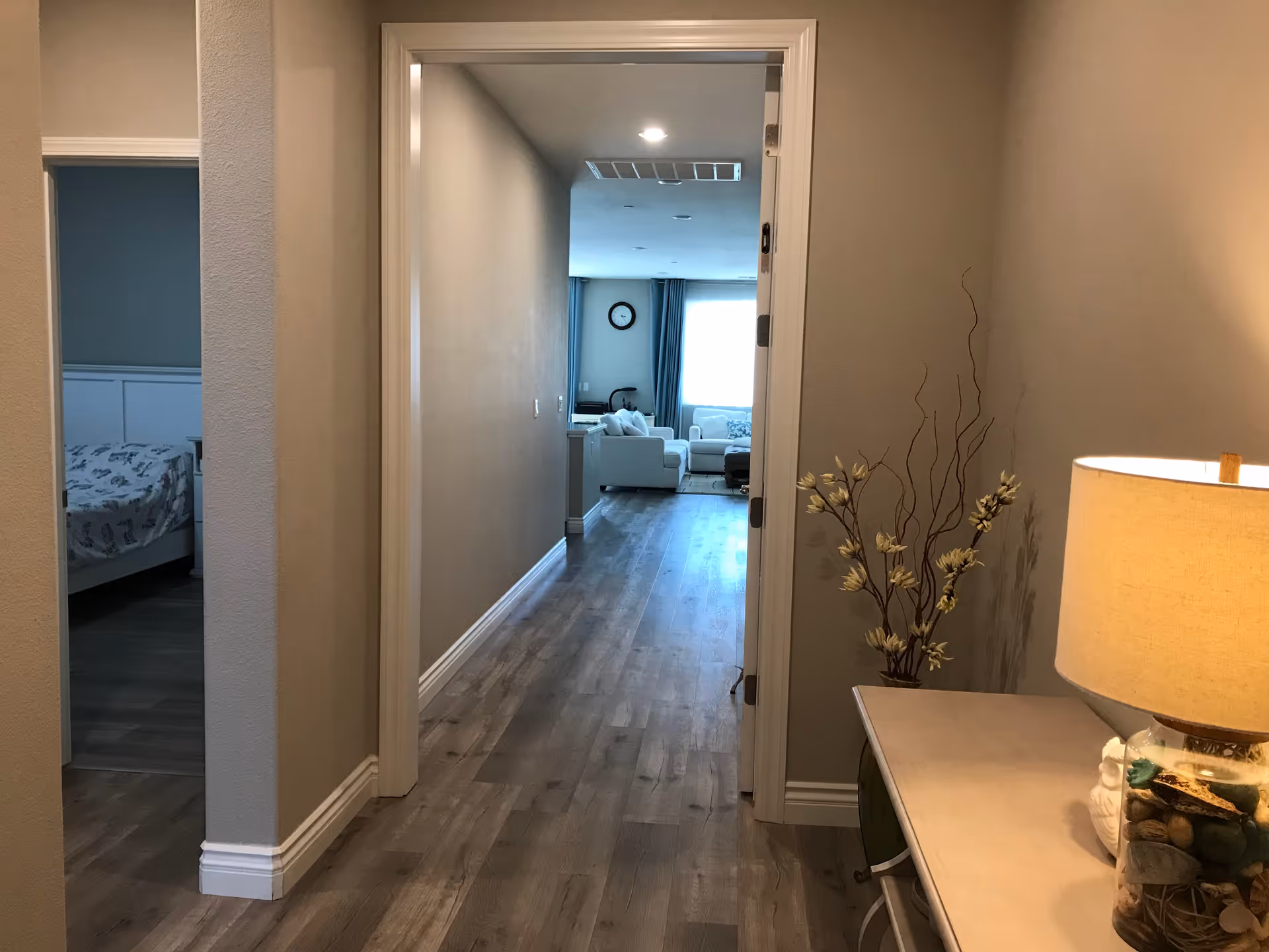 Hallway leading to a bright living room with seating and a clock visible in the distance.
