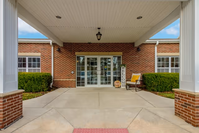 Covered entrance to a brick building with double glass doors. There are two white columns supporting the roof of the entrance area. On the right side near the door, there is a chair with a yellow cushion and a vertical welcome sign. Green bushes flank both sides of the entrance.