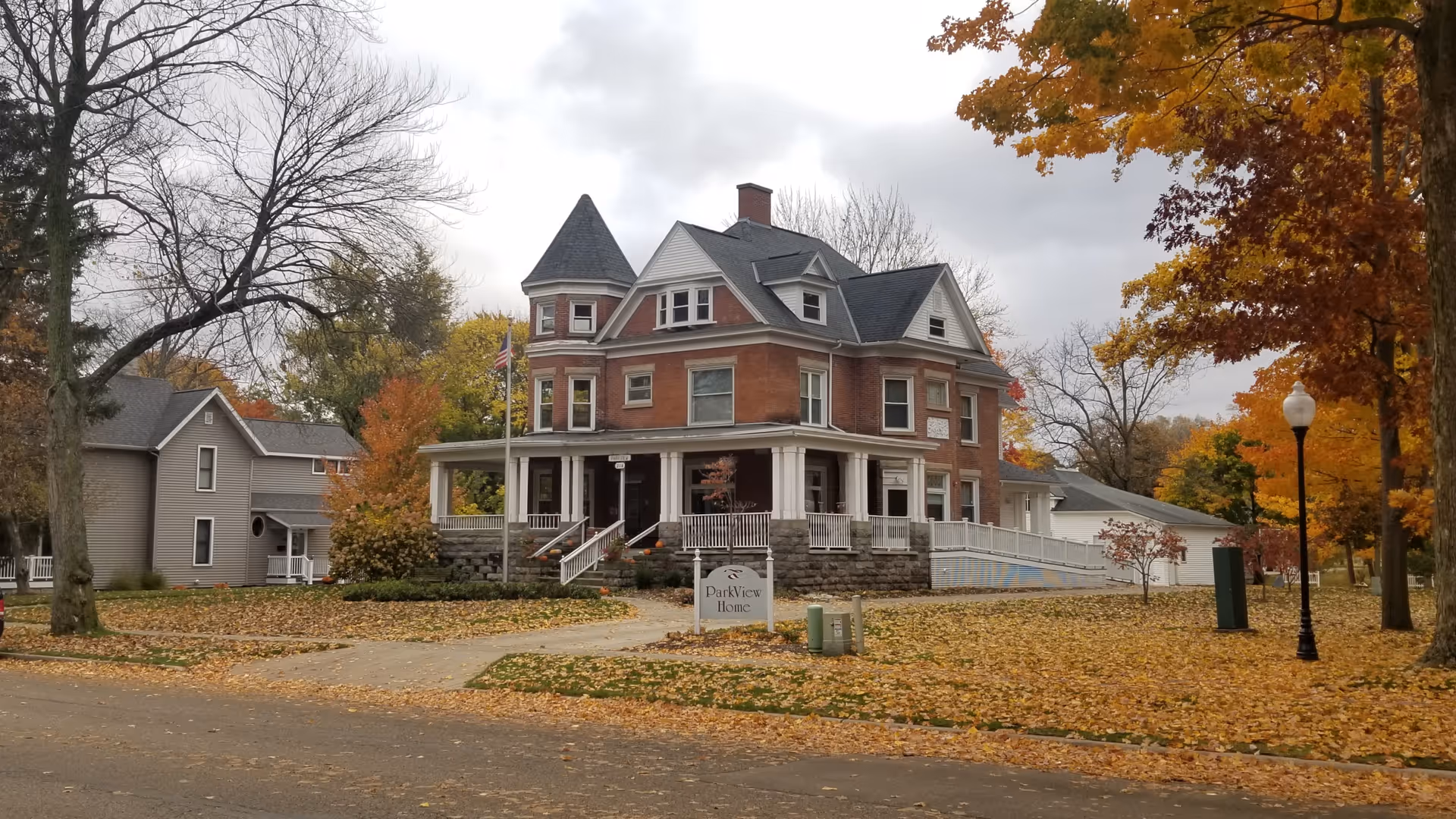 A large brick house with a wraparound porch surrounded by autumn trees.