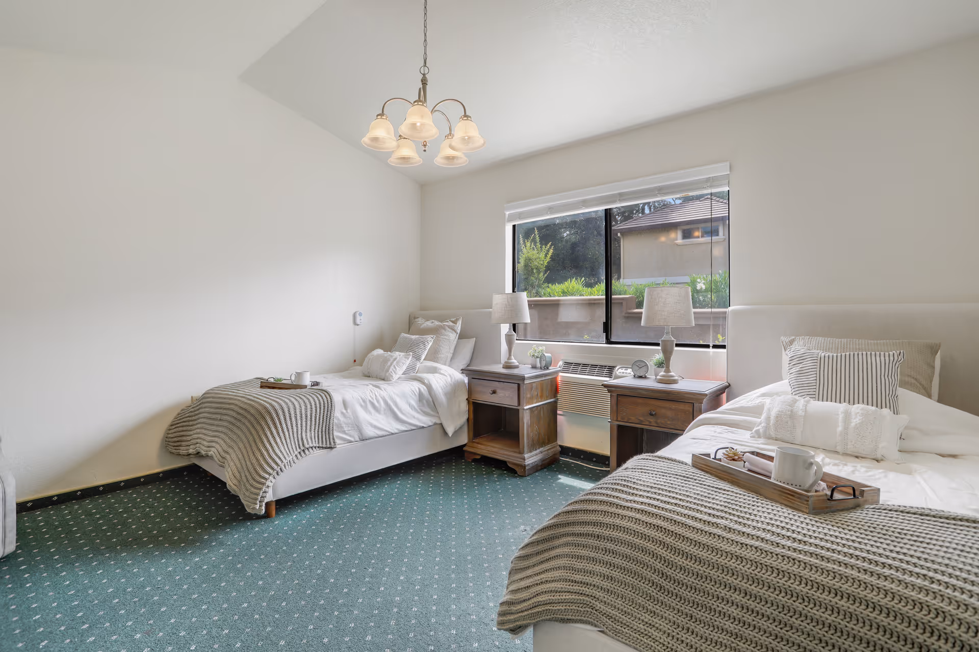 Two single beds in a bright, simply furnished bedroom with nightstands, lamps and a window.