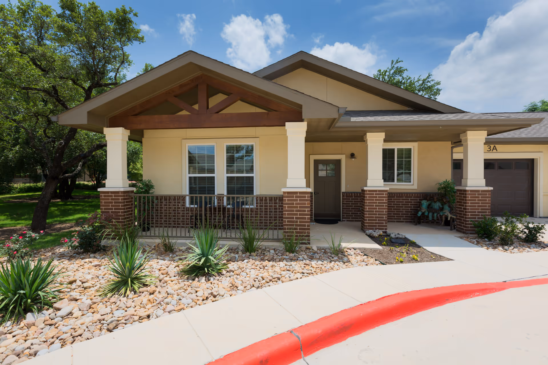 Exterior view of a single-story senior living facility unit with a covered porch supported by brick and white columns. The porch has a railing and two windows, with a door to the right. There are decorative plants and rocks in the landscaped area in front of the porch, and a red-painted curb along the sidewalk. Trees and a partly cloudy sky are visible in the background.