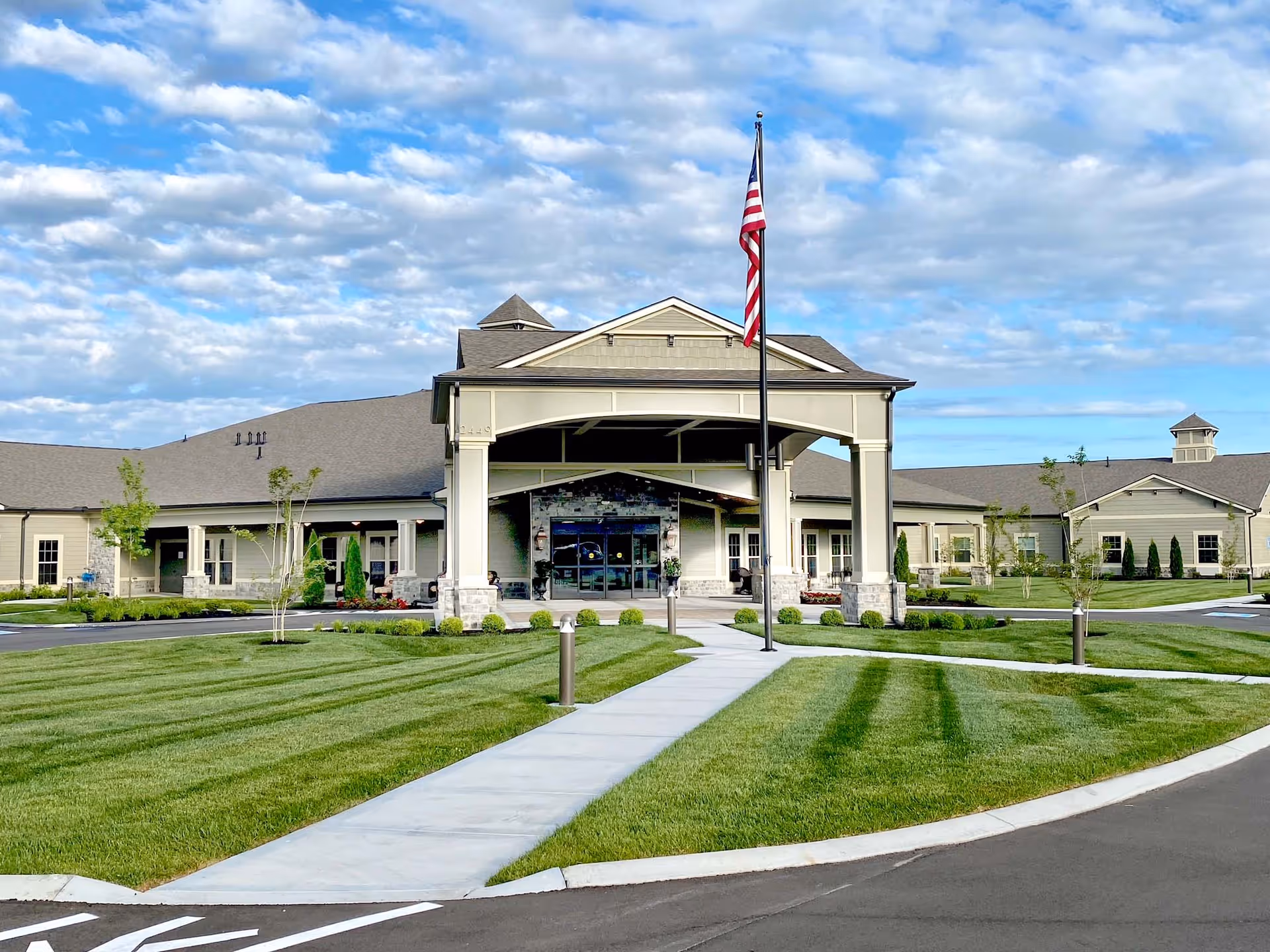 Front entrance of a single-story senior living facility with a covered porte-cochère, an American flag, and manicured lawns.
