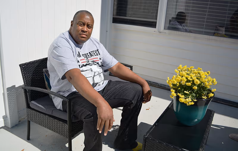 A man wearing a gray t-shirt and black pants is sitting on a black wicker chair on a patio. Next to him is a black wicker table with a green pot containing yellow flowers. The background shows white siding and a window with blinds.