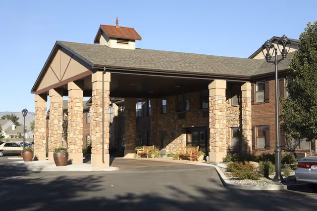 Entrance of a senior living facility with a covered driveway supported by stone and beige pillars, benches near the entrance, and a landscaped area with plants and flowers. The building exterior features stone and brick walls under a clear blue sky.