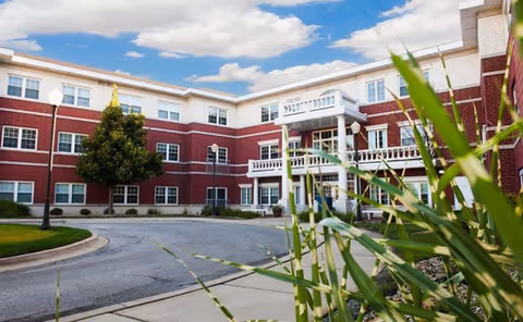 Exterior view of a three-story senior living facility building with red brick and white trim, featuring multiple windows, a balcony, and a driveway with landscaping including green plants and a tree under a partly cloudy sky.